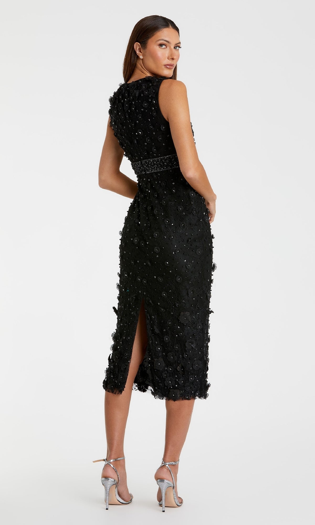 Woman in a sleeveless black Mac Duggal 67853 knee-length party dress with beaded details and back slit, paired with silver high heels, standing against a plain white background.